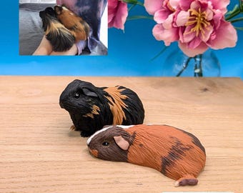 Puede incluir: Dos figuras de cobayas sobre una superficie de madera. Una figura es negra con manchas de color canela, y la otra es marrón, canela y blanca. Una pequeña etiqueta rectangular con el texto Rosie & Cheeky es visible en la esquina inferior derecha.