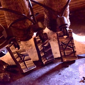 Pode incluir: Três pingentes de prata com cordões de couro e contas de madeira pendurados em um galho de madeira. Cada pingente apresenta um símbolo de runa diferente.