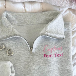 May include: A grey sweatshirt with a pink embroidered design that says "Capricorn" with the text "Font Text" underneath. The sweatshirt has a zip and a mock neck.