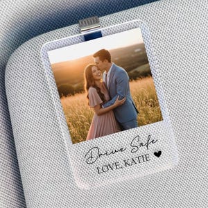 May include: A clear acrylic car visor clip with a photo of a couple in a field at sunset. The clip has the words "Drive Safe, Love, Katie" in script font and a small heart.