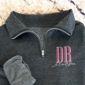 May include: A dark gray quarter-zip sweatshirt with a folded sleeve. The sweatshirt features the pink letters "DR" above the name "Madison" in a script font. The zipper is silver.
