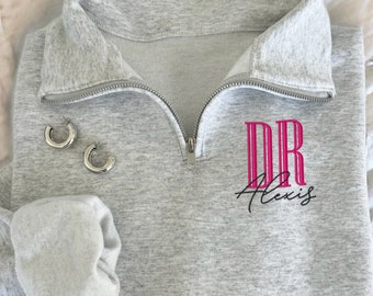 May include: A light gray sweatshirt with a silver zipper and the text "DR Alexis" in pink and black. The sweatshirt has a folded cuff and is paired with silver hoop earrings. The items are arranged on a white, textured surface.