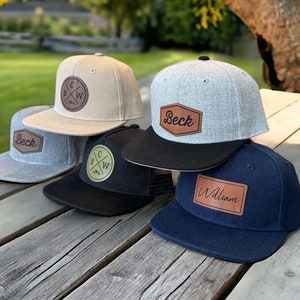 May include: Five baseball caps with different colored fabrics and personalized leather patches. The patches have the names "Beck", "William", and a logo with the letters "B C W" with a mountain graphic.