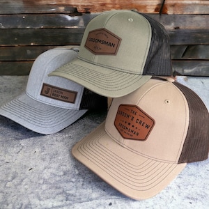 May include: Three baseball caps with mesh backs and leather patches. One is olive green with "Groomsman" on the patch, another is light gray with "Jason Best Man", and the third is tan with "The Groom's Crew".