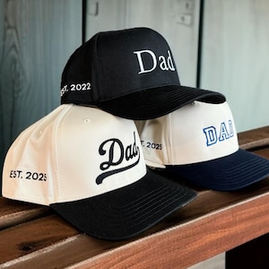 May include: Three baseball caps stacked on a wooden surface. The caps are black, white, and navy blue. Each cap has the word "Dad" embroidered on the front. One cap says "Est. 2022" and another says "Est. 2025".