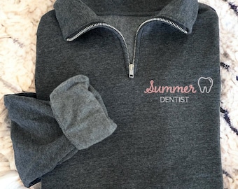 Könnte beinhalten: Ein dunkelgraues Sweatshirt mit Viertelreißverschluss und dem Text "Summer Dentist" in rosa Schrift und einer Zahn-Grafik. Das Sweatshirt hat einen gefalteten Kragen und lange Ärmel. Der Reißverschluss ist silberfarben.