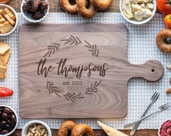 May include: A wooden cutting board with a handle, engraved with the text "The Thompsons est. 2022" in a decorative wreath design. The board is surrounded by various food items, including bagels, tomatoes, nuts, and cheese.