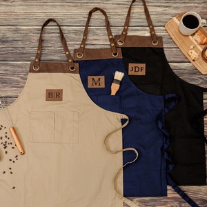 May include: Three brown canvas aprons with leather straps and pockets. Each apron has a personalized leather patch with initials. The aprons are on a wood background.
