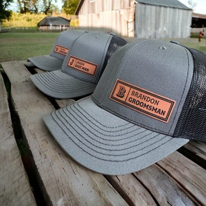 May include: Three gray and black trucker hats with leather patches. The patches have the letters "J", "B", and "G" and the words "Jason Best Man", "Brandon Groomsman", and "Owen Groom".