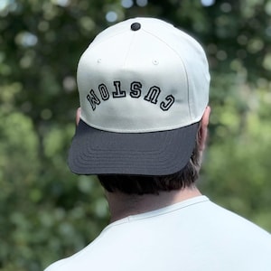 May include: A white baseball cap with a curved brim. The cap has a black brim and the word 'CUSTOM' arched across the front in black letters. The cap is worn by a person, with the back of their head and shoulders visible against a blurred green background.