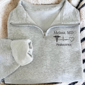 May include: A light gray quarter-zip sweatshirt with the name "Melissa, MD", a medical symbol, a heartbeat line, and a heart, with the word "Pediatrics" below. The sweatshirt is folded on a cream-colored blanket.