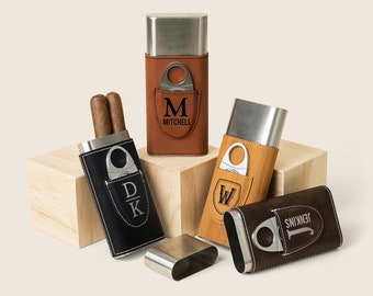 May include: Four personalized leather cigar cases with metal cigar cutters. The cases are brown, black, tan, and dark brown. Each case has a different monogram and name engraved on the front.