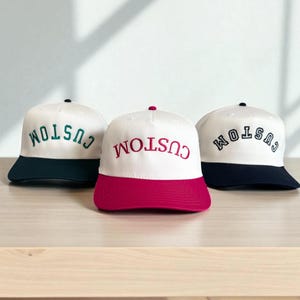 May include: Three white baseball caps with the word "CUSTOM" embroidered in different colors. The caps have colored brims in dark green, red, and navy blue. The caps are displayed on a light brown surface.