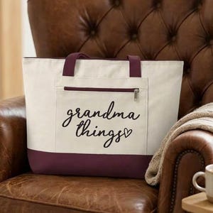 Op de afbeelding: Cr&egrave;mekleurige draagtas met een bordeauxrode basis en handvatten, met een voorvak met ritssluiting. De woorden "grandma things" staan in cursief op de voorkant. De tas staat op een bruine leren fauteuil.
