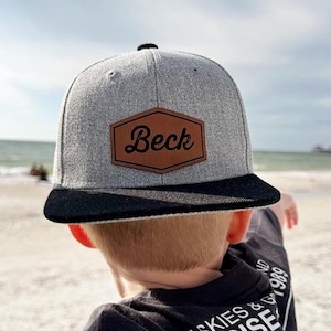 May include: A gray and black baseball cap with a brown leather patch that says "Beck" on it. The cap is worn by a child standing on a beach.