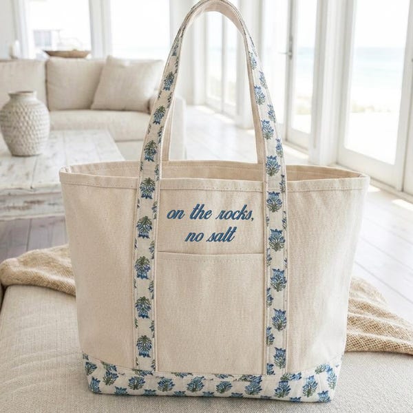 Puede incluir: Bolso tote de lona color crema con ribete y asas florales azules. La bolsa presenta el texto "on the rocks, no salt" en azul. El bolso tote tiene forma rectangular y un bolsillo frontal.