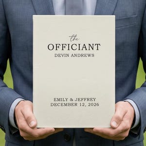 May include: A cream-colored rectangular book cover featuring the black printed text "the OFFICIANT", "DEVIN ANDREWS", and "EMILY & JEFFREY DECEMBER 12, 2026". The cover has a smooth, matte texture and is held by a person wearing a blue suit jacket.