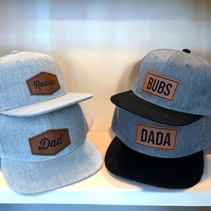 May include: Gray flat-brimmed baseball caps with brown leatherette patches. One stack has "Rowan" and "Dad" on the patches. The other stack has "BUBS" and "DADA" on the patches. The caps have black brims.