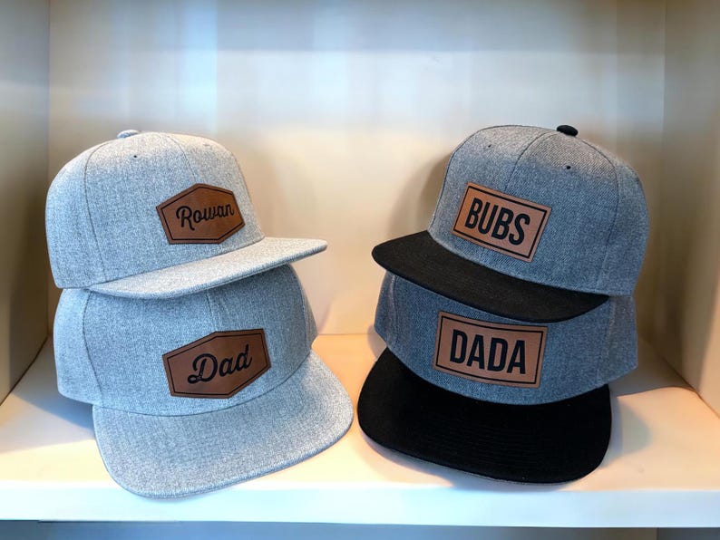 May include: Gray flat-brimmed baseball caps with brown leatherette patches. One stack has "Rowan" and "Dad" on the patches. The other stack has "BUBS" and "DADA" on the patches. The caps have black brims.
