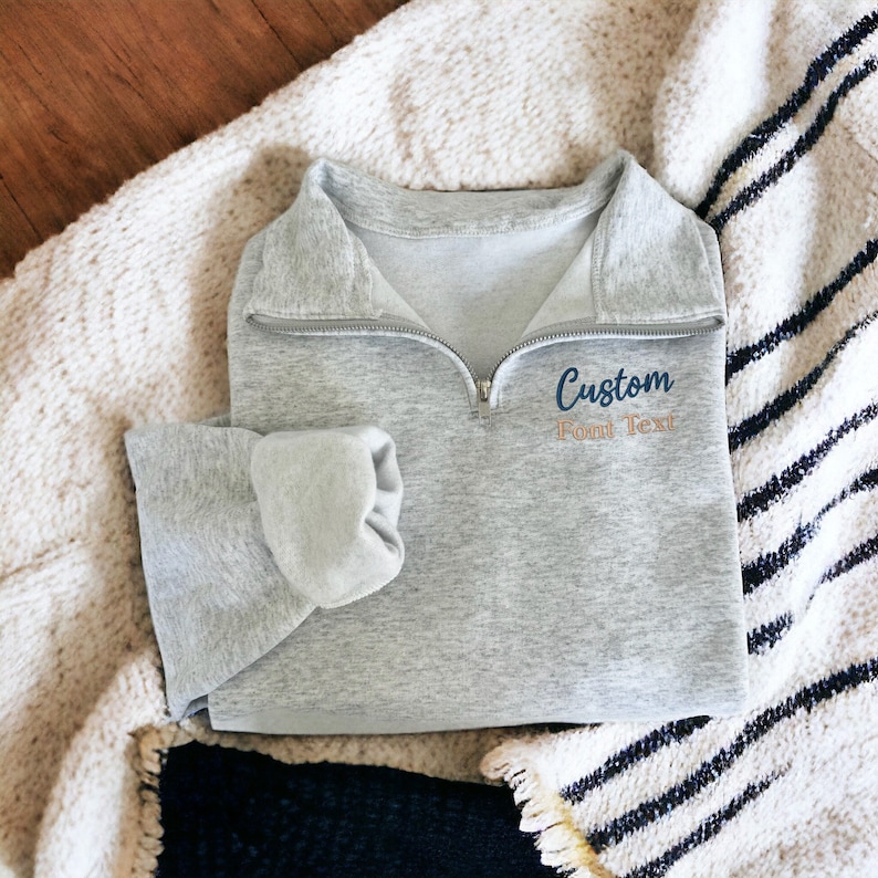 May include: Grey half-zip sweatshirt with a custom embroidered design that says "Custom Font Text" in a navy blue and orange font.