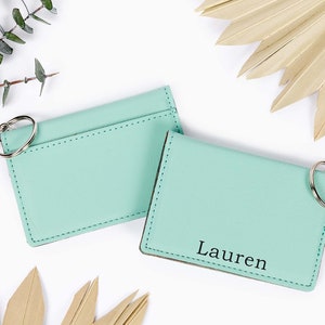 May include: Two mint green cardholders with key rings. One cardholder has the name "Lauren" printed in black. The cardholders are on a white surface with decorative leaves and greenery.