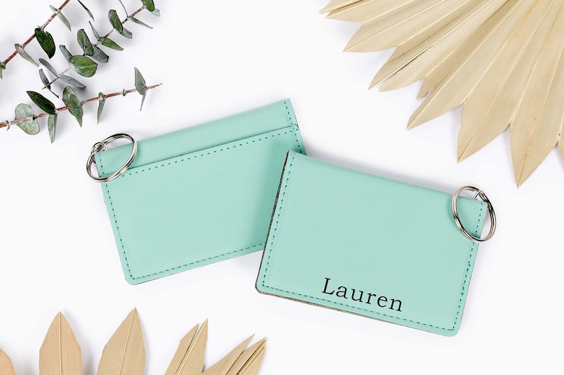 May include: Two mint green cardholders with key rings. One cardholder has the name "Lauren" printed in black. The cardholders are on a white surface with decorative leaves and greenery.