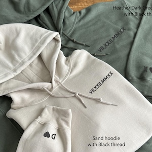 May include: Close-up of two hoodies, one sand-colored and one heather dark green. Both have black thread details, including text and small graphics. The sand hoodie has a drawstring hood and a small heart graphic on the cuff. The green hoodie has an XO heart graphic.