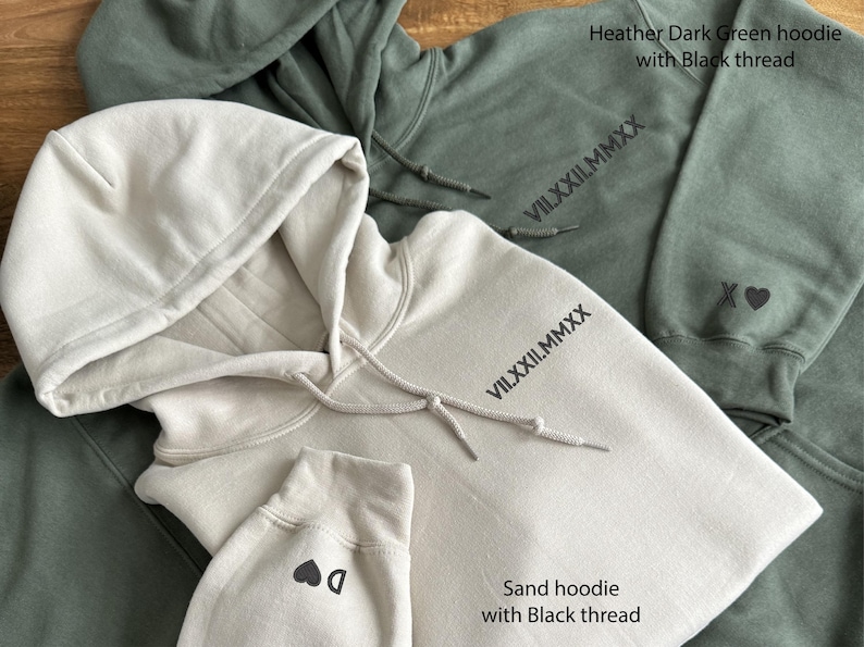 May include: Close-up of two hoodies, one sand-colored and one heather dark green. Both have black thread details, including text and small graphics. The sand hoodie has a drawstring hood and a small heart graphic on the cuff. The green hoodie has an XO heart graphic.