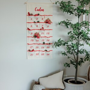 May include: A cream-colored Christmas advent calendar with red accents and the name "Caden" in red script. The calendar has 24 pockets, each numbered and decorated with small gifts and greenery. It hangs from a wooden dowel.