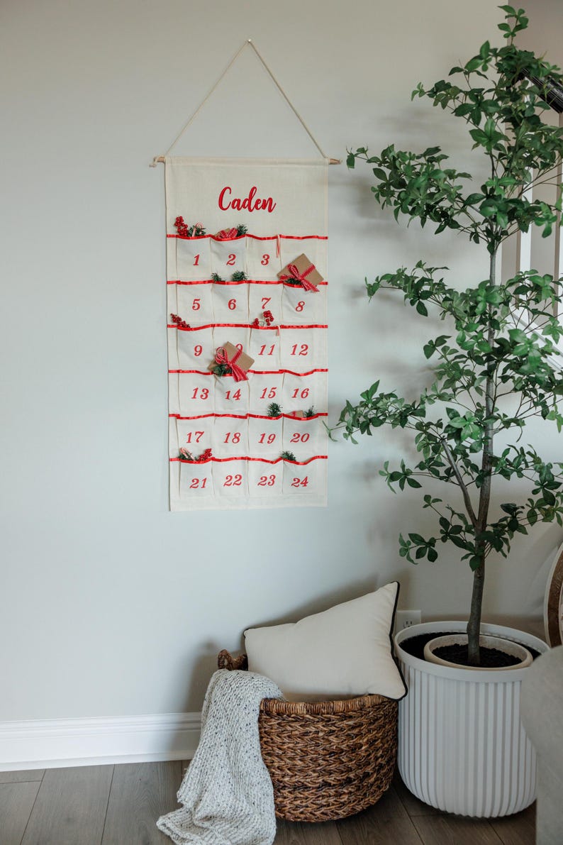 May include: A cream-colored Christmas advent calendar with red accents and the name "Caden" in red script. The calendar has 24 pockets, each numbered and decorated with small gifts and greenery. It hangs from a wooden dowel.