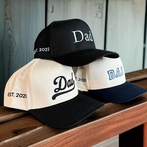 May include: Three baseball caps stacked on a wooden surface. The caps are black, white, and navy blue. Each cap has the word "Dad" embroidered on the front. One cap says "Est. 2022" and another says "Est. 2025".
