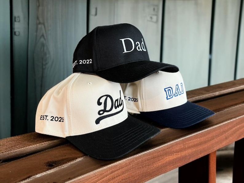May include: Three baseball caps stacked on a wooden surface. The caps are black, white, and navy blue. Each cap has the word "Dad" embroidered on the front. One cap says "Est. 2022" and another says "Est. 2025".