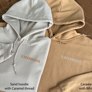 May include: Two hooded sweatshirts, one sand-colored and one caramel-colored, are displayed. The sand hoodie has caramel-colored embroidery and a heart with the letter "S" on the sleeve. The caramel hoodie has white embroidery. Both have drawstring hoods.