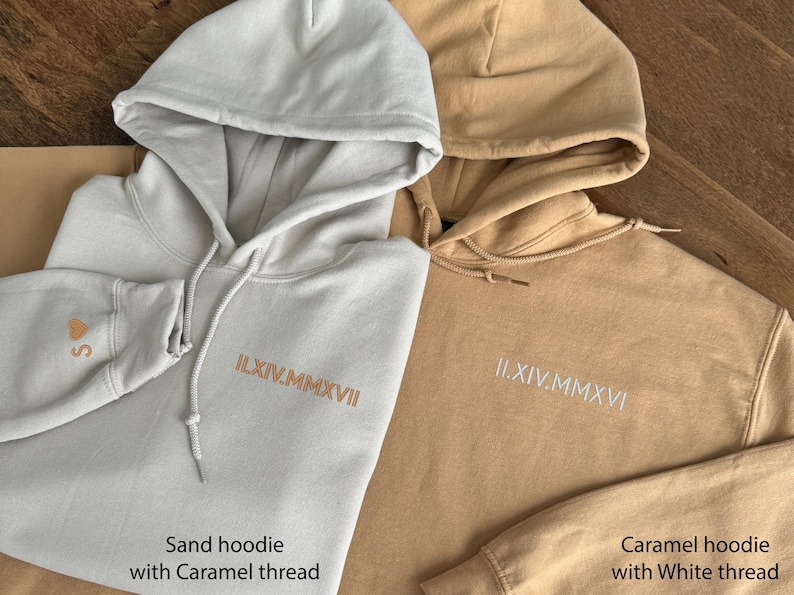 May include: Two hooded sweatshirts, one sand-colored and one caramel-colored, are displayed. The sand hoodie has caramel-colored embroidery and a heart with the letter "S" on the sleeve. The caramel hoodie has white embroidery. Both have drawstring hoods.