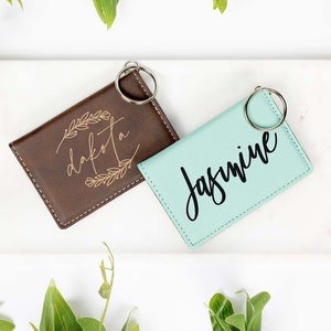 May include: Two personalized keychains. One is brown with "Dakota" in a floral design, the other is mint green with "Jasmine" in cursive. Both have silver key rings. The keychains are on a white surface with green foliage.