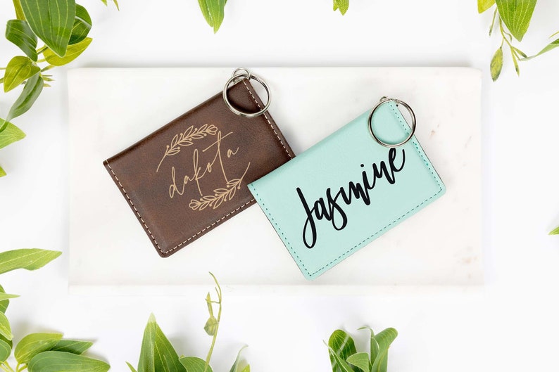 May include: Two personalized keychains. One is brown with "Dakota" in a floral design, the other is mint green with "Jasmine" in cursive. Both have silver key rings. The keychains are on a white surface with green foliage.