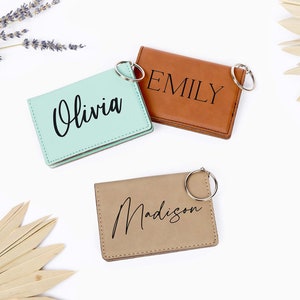 May include: Three personalized keychains in mint green, brown, and tan leather. Each keychain is rectangular and features a name in a stylish script font: "Olivia", "Emily", and "Madison". Silver key rings are attached. Dried lavender and palm fronds are in the background.