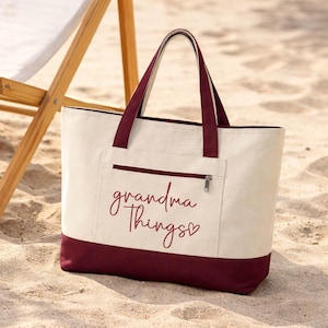 Op de afbeelding: Cr&egrave;mekleurige draagtas met bordeauxrode accenten en handvatten, met de tekst "grandma Things" in een sierlijk lettertype. De tas heeft een ritsvak en ligt op zand.