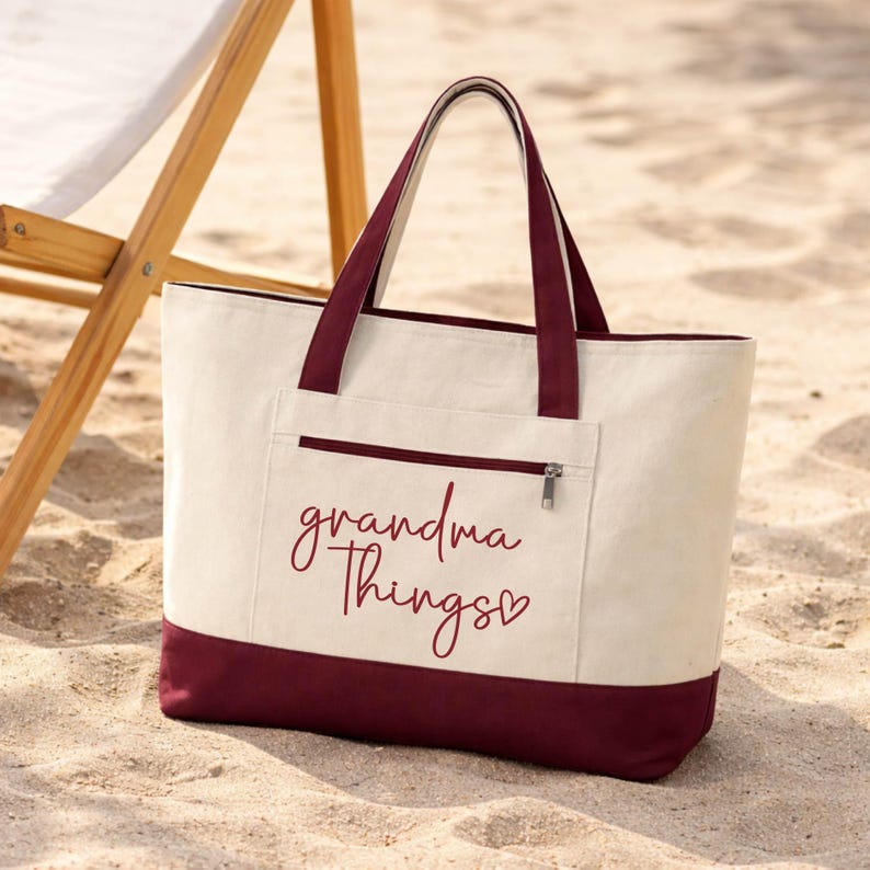 Op de afbeelding: Cr&egrave;mekleurige draagtas met bordeauxrode accenten en handvatten, met de tekst "grandma Things" in een sierlijk lettertype. De tas heeft een ritsvak en ligt op zand.