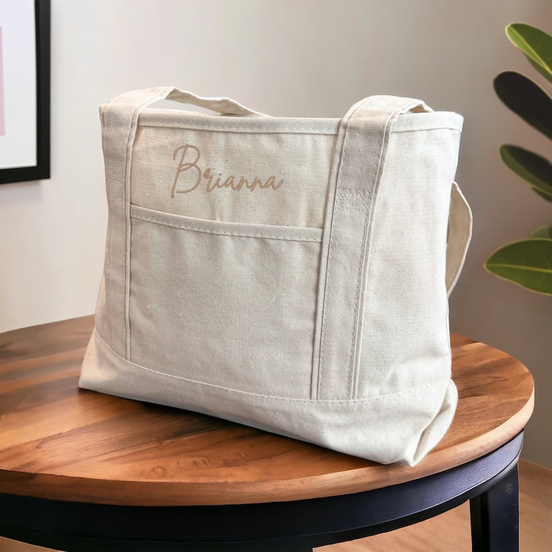 May include: A beige canvas tote bag with a large pocket on the front. The bag has two handles and the name "Brianna" is embroidered on the front in a light brown color.