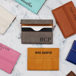 May include: Assortment of personalized leather card holders in various colors, including pink, blue, brown, gray, and beige. Each holder features a stitched design and a personalized name or initials.