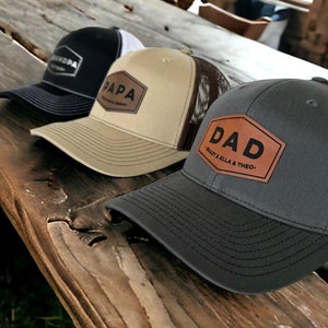 May include: Three trucker hats with brown leather patches. The patches have the words "Grandpa", "Papa", and "Dad" on them. The hats are on a wooden surface.