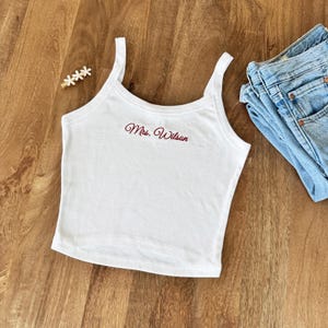 May include: White tank top with the embroidered text "Mrs. Wilson" in a cursive font. The top has thin straps and a scoop neckline. A pair of blue jeans is visible in the background.