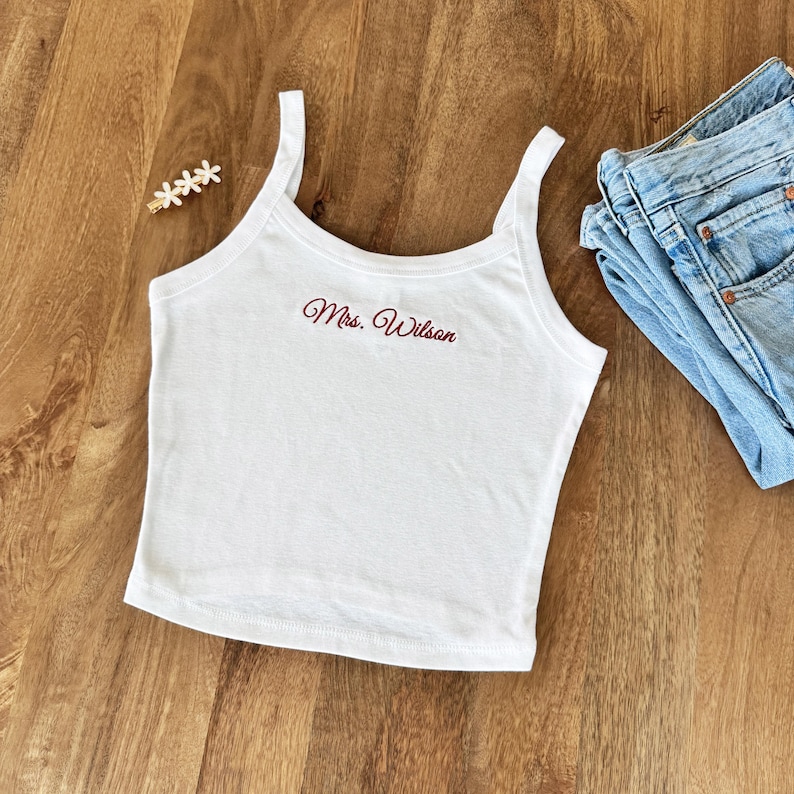 May include: White tank top with the embroidered text "Mrs. Wilson" in a cursive font. The top has thin straps and a scoop neckline. A pair of blue jeans is visible in the background.
