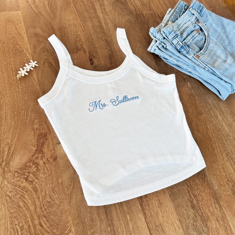 May include: White tank top with the embroidered text "Mrs. Sullivan" in light blue. The top has thin straps and a scoop neckline. A pair of blue jeans and a floral hair clip are also in the image.