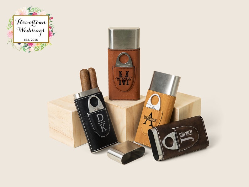 May include: Four personalized cigar cases with a built-in cutter. The cases are made of metal and leather and come in brown, black, and tan. The cases are engraved with initials and names, including "Mitchell", "Harrison", "Jenkins", and "DK".