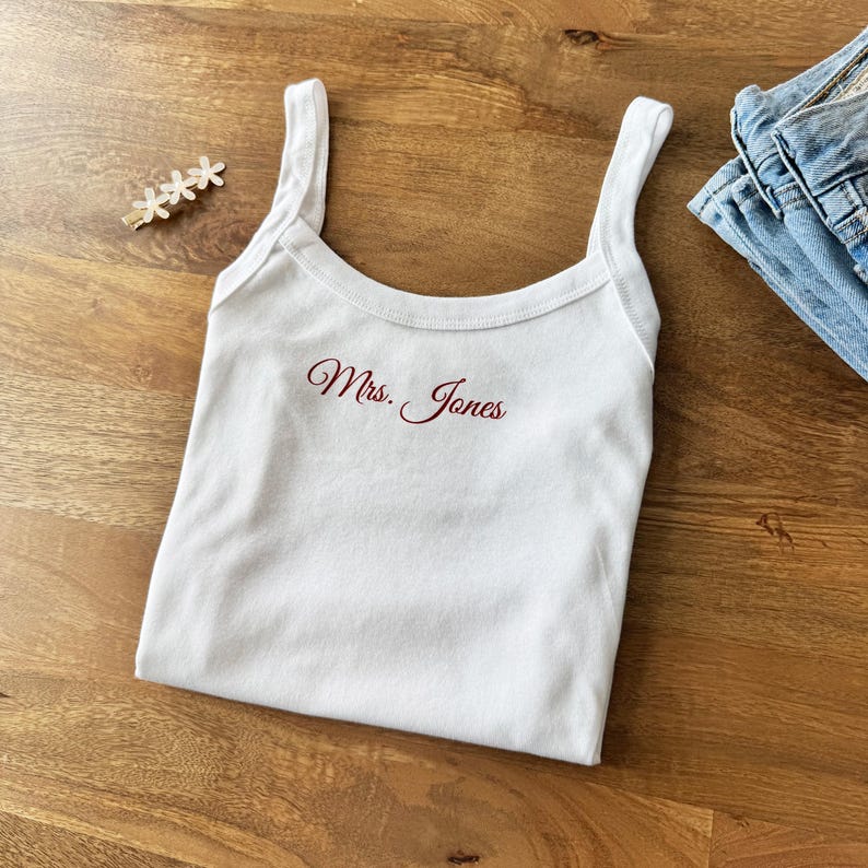 May include: White tank top with thin straps, featuring the embroidered text "Mrs. Jones" in a burgundy script. The top is folded on a wooden surface, with a hair clip and a pair of blue jeans in the background.