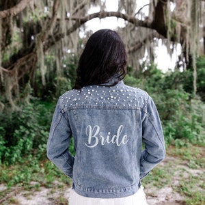 May include: A woman wearing a white dress and a blue denim jacket with pearl embellishments and the word "Bride" written on the back.
