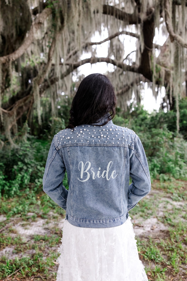 May include: A woman wearing a white dress and a blue denim jacket with pearl embellishments and the word "Bride" written on the back.