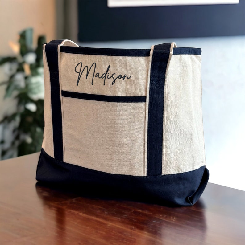 May include: A beige canvas tote bag with black trim and handles. The bag has a large pocket on the front with the name "Madison" embroidered in black.